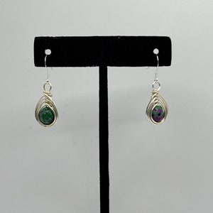May include: A pair of silver wire wrapped earrings with green and pink gemstones. The earrings are displayed on a black stand.