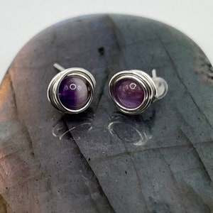 May include: A pair of amethyst gemstone earrings with silver wire wrapping. The earrings are displayed on a gray stone surface.