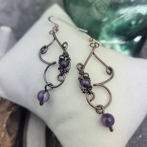 May include: Artisan-crafted earrings with a unique wire design. The earrings feature a combination of copper and dark metal, with small, round, purple amethyst beads. The earrings are displayed on a white pillow.