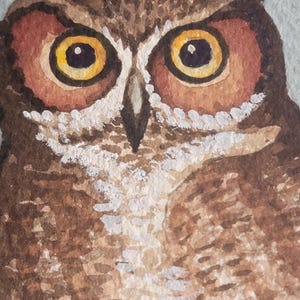 May include: Watercolor painting of an owl's face, featuring large, round yellow eyes with black pupils. The owl's feathers are depicted in shades of brown, with white accents around the eyes and a light gray background. The artwork showcases detailed brushstrokes.