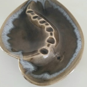 May include: A brown ceramic ashtray with a white glaze around the edge. The ashtray is shaped like a leaf with a raised design in the center.