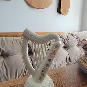 May include: A white ceramic harp figurine with intricate embossed floral and Celtic knot designs. The harp has a decorative base and is displayed on a wooden surface. The harp's strings are visible, and the design includes copper-colored accents.