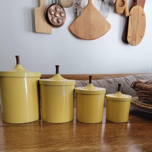 May include: Four yellow canisters with dark brown wooden finials. The canisters are arranged in a row, decreasing in size from left to right. The canisters are on a wooden table. Kitchen decor.