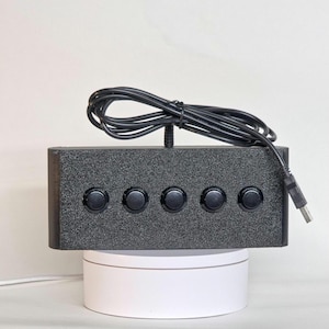 May include: A rectangular black control box with a textured finish featuring five circular black push buttons arranged in a horizontal row. A black USB cable is attached to the top center of the device, coiled neatly above the unit.