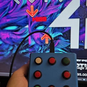 Puede incluir: Un controlador azul con nueve botones de colores, sostenido frente a una pantalla de ordenador que muestra un juego de ritmo. La pantalla muestra flechas y la palabra "MISS". Los botones son rojos, verdes, negros y blancos.