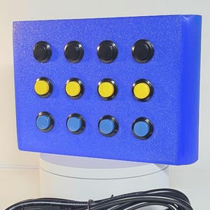 May include: A rectangular blue controller box featuring twelve circular push buttons arranged in three rows of four. The top row has black buttons, the middle row has yellow buttons, and the bottom row has blue buttons. A black USB cable is included.