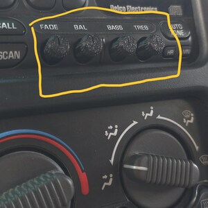 May include: A set of four black textured control knobs on a Delco Electronics car radio interface. The knobs are labeled FADE, BAL, BASS, and TREB, positioned above the climate control dials featuring A/C, fan, and defrost icons in a vehicle.