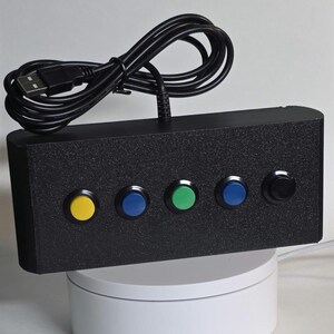 May include: A rectangular black controller box with a textured finish featuring five circular push buttons in yellow, blue, green, blue, and black. A black USB cable is attached to the top of the device, which is positioned on a white base.