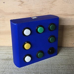May include: A rectangular, cobalt blue device with nine circular buttons of various colors: white, green, yellow, and black. The buttons are arranged in a 3x3 grid on the front of the device. The device has a small rectangular opening on the top.
