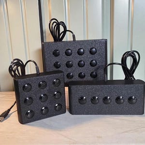 May include: Three black rectangular macro keypad controllers with textured surfaces and black circular buttons. One unit features a three-by-three grid, another a three-by-four grid, and the third a single row of five buttons, each with a black USB cable.