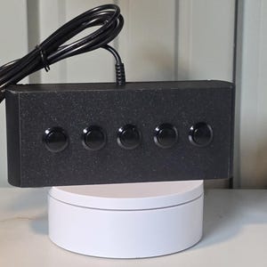 May include: A rectangular black control box with a textured finish featuring five circular black push buttons arranged in a horizontal row. A black electrical cord extends from the top center of the unit, which is positioned on a white base.