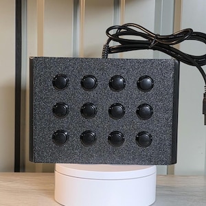 May include: A rectangular black controller box with a textured, glittery finish featuring twelve circular black push buttons arranged in a three-by-four grid. A black power cord extends from the top center of the device, which is positioned upright.