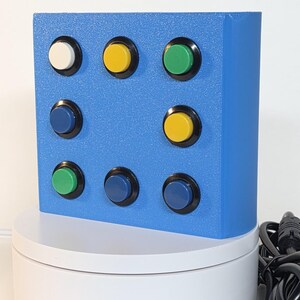 May include: A blue rectangular controller box featuring eight circular push buttons arranged in a grid pattern. The buttons are colored white, yellow, green, and blue, each set within a black circular frame. A black power cord is attached to the side.