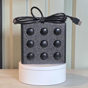 May include: A square black control box with a textured, glittery finish featuring a three-by-three grid of nine circular black push buttons. A black USB cable is coiled on top of the device, extending to the right with a standard USB connector.