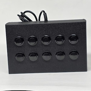 May include: A rectangular black control box featuring ten circular black push buttons arranged in two horizontal rows of five. A black electrical cord extends from the top edge of the device, which has a textured matte finish.