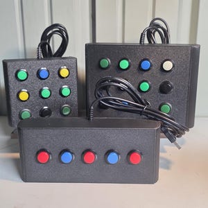 May include: Three black rectangular control boxes with wired USB connections. One box features a 3x3 grid of green, blue, yellow, and black buttons. Another has a 2x4 grid of green, blue, and black buttons. The third is a horizontal row of five buttons.