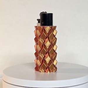 May include: A standard black lighter encased in a 3D-printed sleeve featuring a repeating geometric diamond pattern. The sleeve displays a metallic gradient finish that transitions between shades of copper, bronze, and gold across its surface.