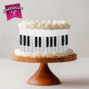 May include: A round cake decorated to resemble a piano keyboard, with black and white keys. The cake is topped with white frosting swirls and sits on a wooden cake stand. The logo "Cakesalot" is in the upper left corner.