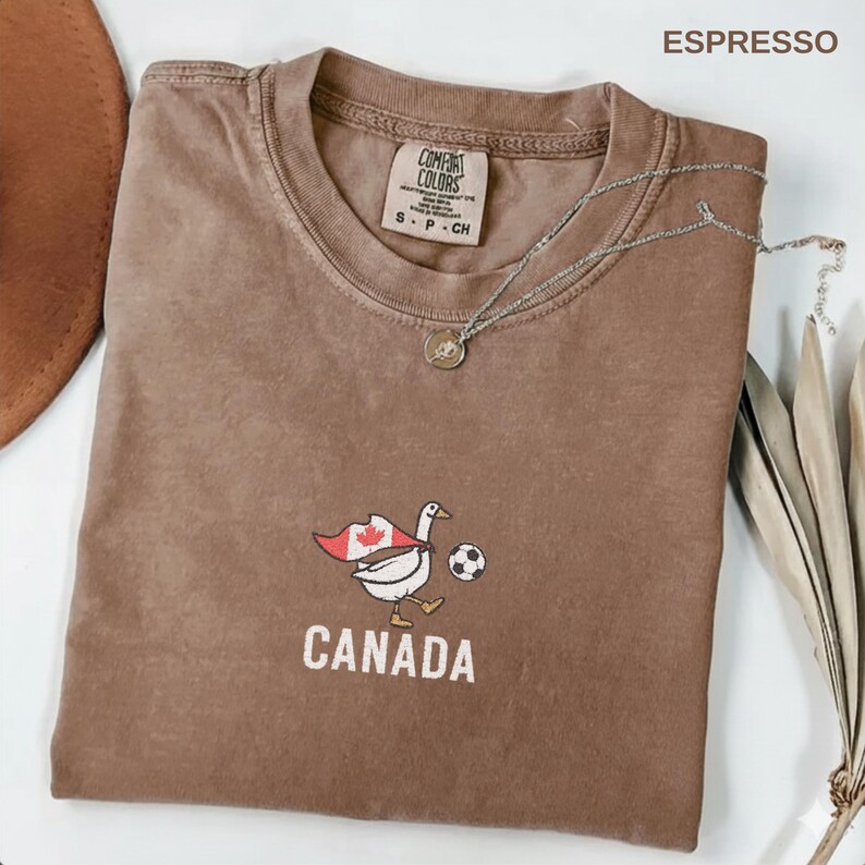 以下が含まれることがあります： エスプレッソ色のTシャツ。カナダの国旗のマントを着てサッカーをするガチョウのグラフィックと、下に「CANADA」の文字。シャツにはComfort Colorsのタグとシルバーのネックレスが付いています。白い面に折りたたまれています。