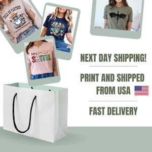May include: A white shopping bag with black handles is in the foreground. Several t-shirts with graphic designs are displayed. The shirts have text and illustrations. Text on the shirts includes "HOLD ON PARTNER", "I'm Overstimulated", and "Mrs. SMITH".