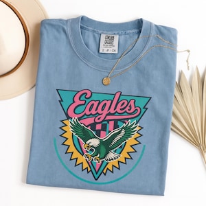 May include: A light blue t-shirt featuring a graphic of an eagle with the word "Eagles" in pink and teal. The shirt has a crew neck and short sleeves. A gold necklace and a hat are also visible.