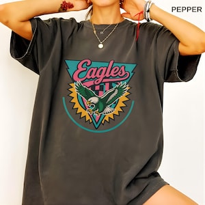 May include: Dark grey t-shirt featuring a colorful graphic of an eagle with outstretched wings, the word "Eagles" in pink and teal, and a teal and yellow design. The shirt is a relaxed fit.