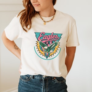 May include: Cream-colored t-shirt featuring a colorful Eagles logo. The logo includes the word "Eagles" in pink script, a green and white eagle, and a teal and yellow design.
