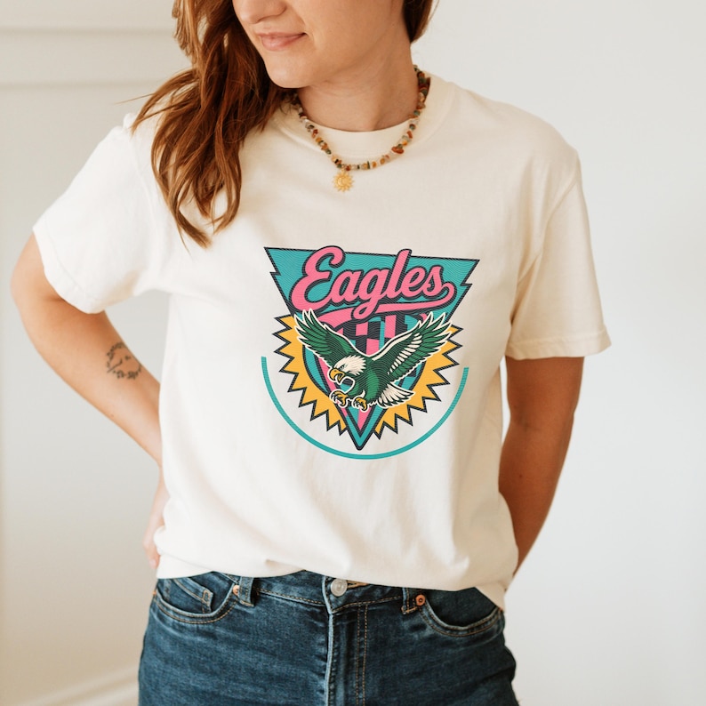 May include: Cream-colored t-shirt featuring a colorful Eagles logo. The logo includes the word "Eagles" in pink script, a green and white eagle, and a teal and yellow design.