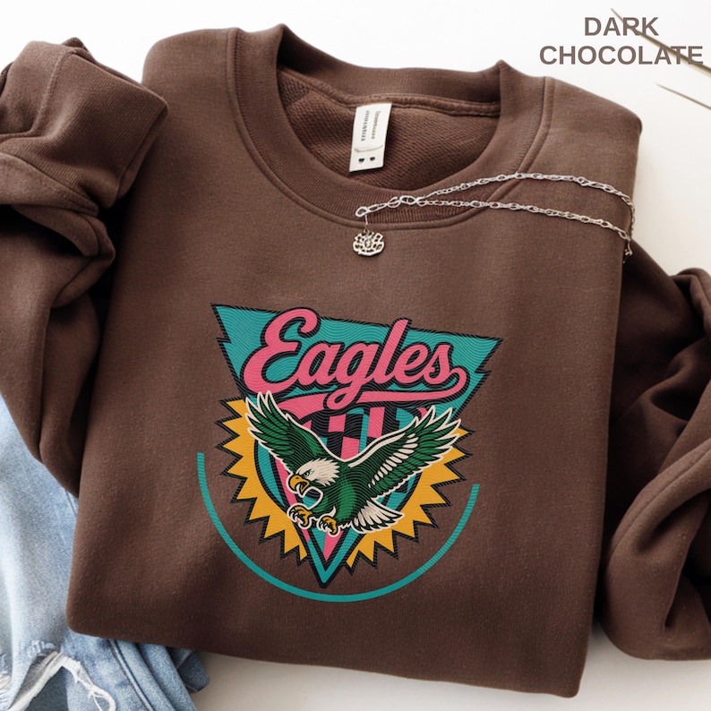 May include: Dark chocolate-colored sweatshirt with a colorful Eagles graphic. The graphic features the word "Eagles" in pink, a teal and yellow design, and a green and white eagle. A silver necklace with a small pendant is draped over the sweatshirt.