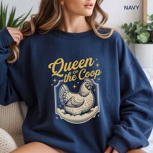 May include: Navy blue sweatshirt with the text "Queen of the Coop" in gold script above a cream-colored hen illustration. The hen is inside a decorative frame with star accents. The word "NAVY" is printed in the upper right corner.