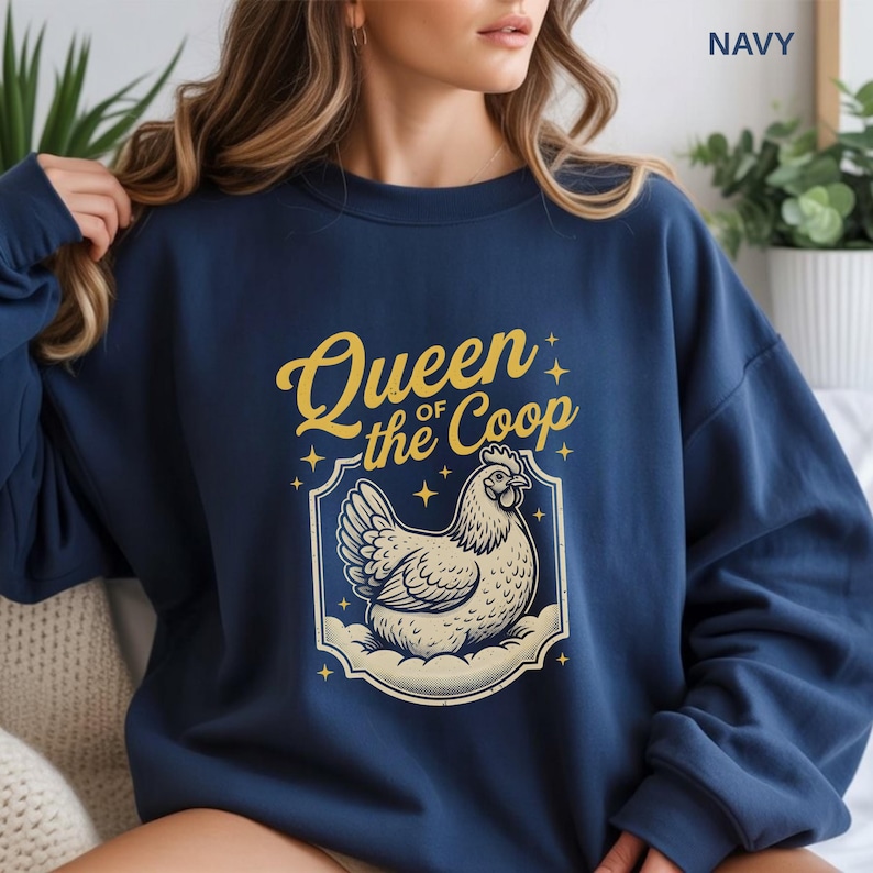 May include: Navy blue sweatshirt with the text "Queen of the Coop" in gold script above a cream-colored hen illustration. The hen is inside a decorative frame with star accents. The word "NAVY" is printed in the upper right corner.