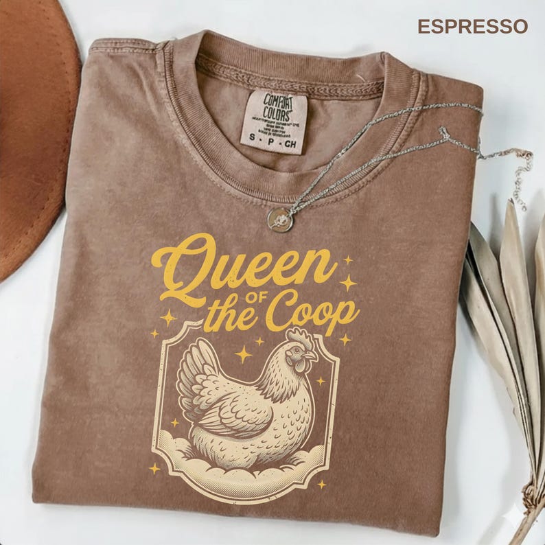 May include: Espresso-colored t-shirt with the text "Queen of the Coop" in yellow, featuring a hen illustration. The shirt has a crew neck and short sleeves. A silver necklace with a pendant is draped across the shirt.