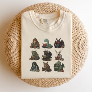 Op de afbeelding: Crèmekleurig T-shirt met negen cartoonmonsters die boeken lezen. De monsters zijn onder meer een Bigfoot, een Loch Ness-monster, een motachtig wezen, een draak, een geitachtige demon, een hertachtig wezen, een geest, een vosachtig wezen en een Cthulhu-achtig monster.