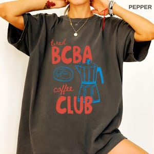 Pode incluir: T-shirt cinza escuro com as palavras "tired BCBA coffee CLUB" em vermelho, e uma ilustração azul de uma cafeteira e um bolo. A camisa tem um corte folgado.