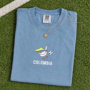 May include: A dusty blue t-shirt featuring a graphic of a white goose wearing a cape with the yellow, blue, and red colors of the Colombian flag, kicking a soccer ball. The word COLOMBIA is printed in white below the goose. A gold necklace is present.
