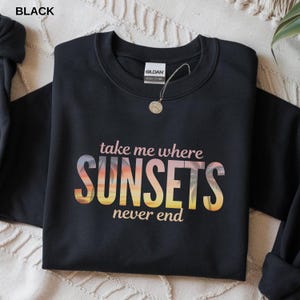 Könnte beinhalten: Schwarzes Sweatshirt mit dem Text "take me where SUNSETS never end" in einer Schriftart im Sonnenuntergangs-Design. Das Sweatshirt hat einen Rundhalsausschnitt und lange Ärmel. Eine goldene Halskette hängt am Kragen.