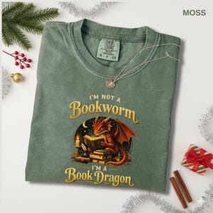 May include: Moss green t-shirt with the text "I'm Not a Bookworm, I'm a Book Dragon" and a graphic of a red dragon reading a book. The shirt has a crew neck and short sleeves. A silver necklace is draped across the shirt.