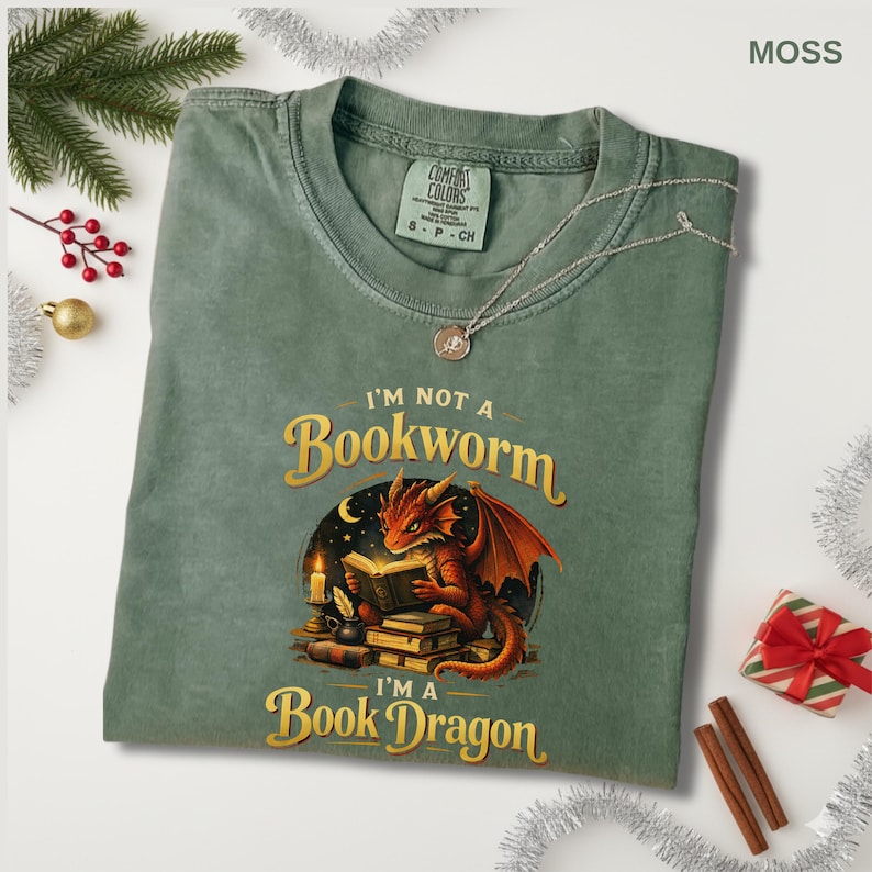 May include: Moss green t-shirt with the text "I'm Not a Bookworm, I'm a Book Dragon" and a graphic of a red dragon reading a book. The shirt has a crew neck and short sleeves. A silver necklace is draped across the shirt.