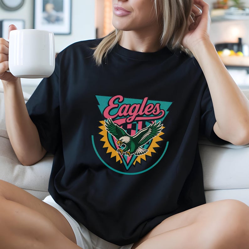 May include: Black t-shirt featuring a graphic of an eagle with outstretched wings, set within a teal and yellow design. The word "Eagles" is arched above the eagle in pink and teal. A classic crew neck tee.