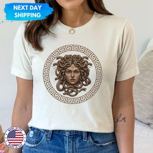 Può includere: Una maglietta bianca con una grafica marrone della testa di Medusa circondata da serpenti. La grafica è all'interno di un cerchio marrone con un motivo a chiave greca.