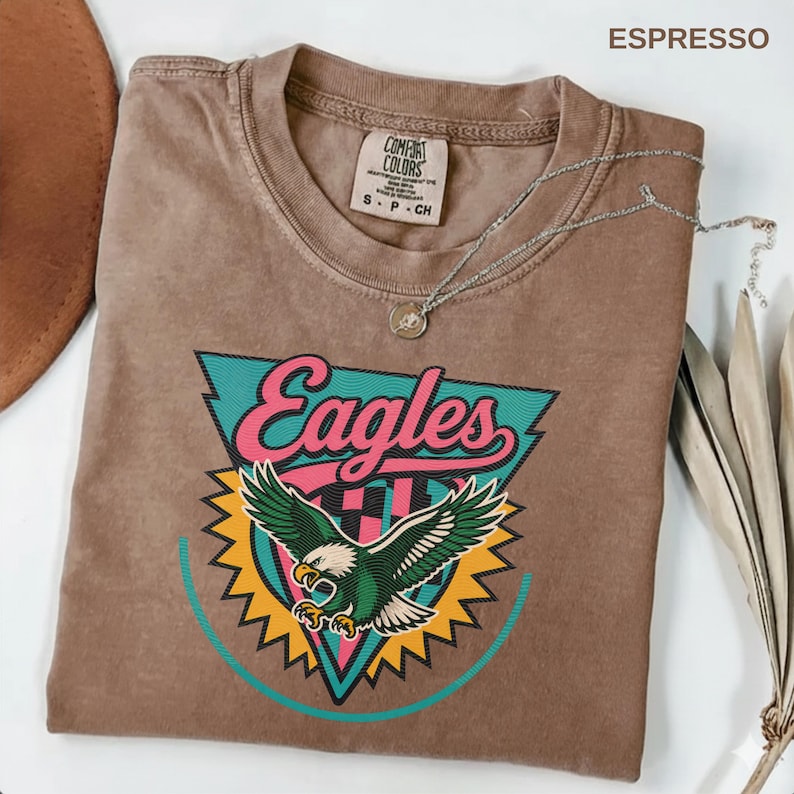 May include: Espresso-colored t-shirt with an "Eagles" graphic. The graphic features a green and white eagle with a pink and teal triangle design. A silver necklace with a pendant is draped across the shirt.