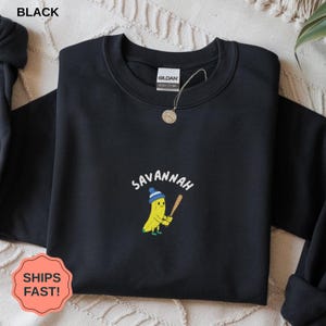 Könnte beinhalten: Schwarzes Sweatshirt mit dem Wort "Savannah" über einer Cartoon-Banane, die eine blau-weiße Mütze trägt und einen Baseballschläger hält. Das Sweatshirt hat einen Rundhalsausschnitt und ein "Gildan"-Etikett.
