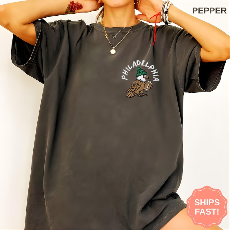 May include: Dark gray t-shirt with a graphic of an eagle wearing a green beanie and holding a football. The word "PHILADELPHIA" is arched above the eagle in white lettering. The shirt has a relaxed fit.
