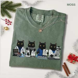May include: Moss green t-shirt with a graphic of four black cats dressed as optometrists. The cats are wearing lab coats and holding medical tools. A silver necklace with a pendant is draped on the shirt.