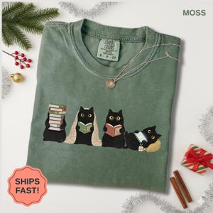 May include: Moss green Comfort Colors t-shirt featuring a graphic of four black cats reading books. One cat holds a stack of books, another is under a blanket, and one wears glasses. A silver necklace and the text "SHIPS FAST!" are also visible.