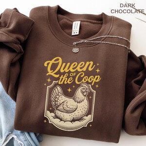 May include: Dark chocolate-colored sweatshirt with the text "Queen of the Coop" in gold lettering, featuring a detailed illustration of a hen. A silver necklace with a pendant is draped across the sweatshirt.