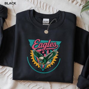 May include: Black crewneck sweatshirt with a colorful graphic. The graphic features the word "Eagles" in pink script above an eagle with green and white feathers. The eagle is set against a teal triangle and yellow sunburst.