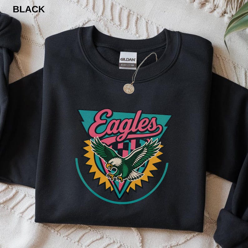 May include: Black crewneck sweatshirt with a colorful graphic. The graphic features the word "Eagles" in pink script above an eagle with green and white feathers. The eagle is set against a teal triangle and yellow sunburst.