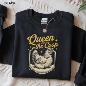 May include: Black sweatshirt with the text "Queen of the Coop" in gold script above a cream-colored illustration of a hen. The design is framed by a decorative border with star accents. The word "BLACK" is in the top left corner.