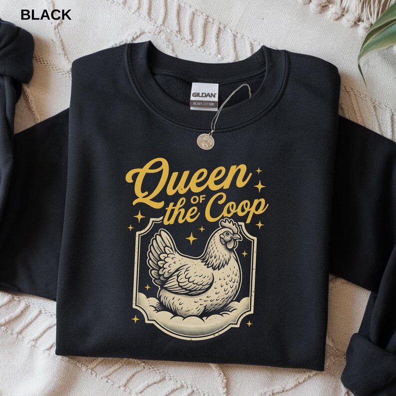 May include: Black sweatshirt with the text "Queen of the Coop" in gold script above a cream-colored illustration of a hen. The design is framed by a decorative border with star accents. The word "BLACK" is in the top left corner.
